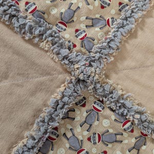 May include: A rag quilt featuring a repeating pattern of gray sock monkeys with red hats and white accents on a tan background. The quilt is constructed with exposed, frayed gray fabric seams that create a textured, cross-hatched grid pattern.