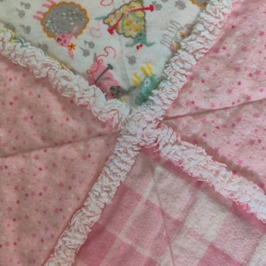 May include: A pink and white baby blanket with a white ruffled edge. The blanket is made of soft, plush fabric and features a variety of patterns, including stars, gingham, and a whimsical animal print.