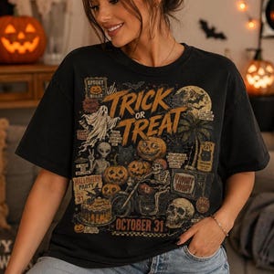May include: A black t-shirt featuring a detailed graphic print with the text "TRICK OR TREAT" and "OCTOBER 31". The design includes illustrations of a ghost, an alien, a skeleton riding a motorcycle, pumpkins, a skull, and a cake.