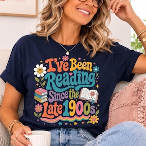 May include: A navy blue t-shirt featuring a colourful graphic with the text I've Been Reading Since the Late 1900s. The design includes illustrations of stacked books, an open book, flowers, and stars in shades of yellow, pink, and teal.