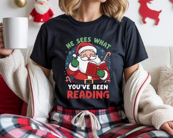Op de afbeelding: Zwart t-shirt met een afbeelding van de kerstman die een boek leest, omringd door de tekst "He sees what you've been reading". De kerstman-illustratie staat in een cirkel met sneeuwvlokken en een zuurstok.