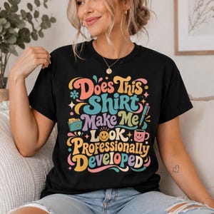 May include: A black short-sleeved t-shirt featuring a colorful graphic with the text Does This Shirt Make Me Look Professionally Developed. The design includes illustrations of a folder, a pen, a coffee mug, and a smiley face in retro colors.