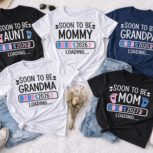 May include: Five t-shirts with pregnancy announcement graphics. The shirts are black, white, and navy blue, with text that reads "Soon to be Aunt", "Mommy", "Grandpa", "Grandma", and "Mom". Each shirt has a loading bar graphic and the year 2026 or 2027.