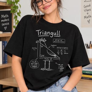 May include: A black short-sleeved t-shirt featuring a white graphic of a seagull wearing glasses, labeled Triangull. The design includes geometric math formulas, a 30-60-90 triangle diagram, and text reading I've got great angles and coastal confidence.