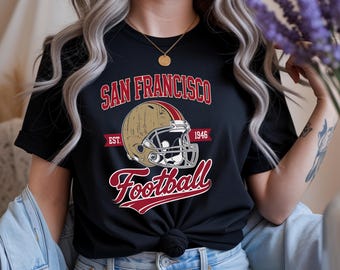 May include: Black t-shirt with a football helmet graphic and the text "SAN FRANCISCO Football" in red and white. The design includes the year "1946" and is a vintage-style design.