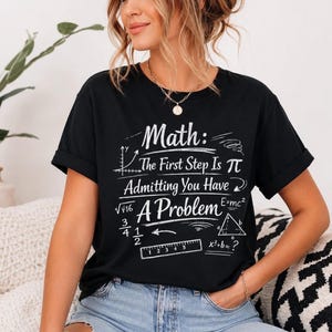 May include: A black short-sleeved t-shirt featuring white text that reads: Math: The First Step Is Admitting You Have A Problem. The design includes various mathematical symbols, equations, a triangle, and a ruler illustration printed on the front.
