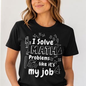 May include: A black short-sleeved t-shirt featuring white graphic text that reads I Solve MATH Problems like it's my job. The design includes various mathematical symbols, a ruler, a triangle, a calculator, and a cone shape printed on the front.
