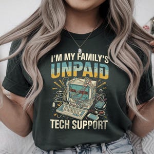 Może przedstawiać: Ciemnozielona koszulka z napisem "I'M MY FAMILY'S UNPAID TECH SUPPORT". Grafika przedstawia komputer w stylu vintage z przewodami, klawiaturą i napisem "SERIAL ERROR" na ekranie. Projekt ma estetykę retro i vintage.