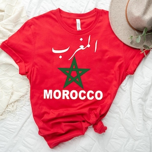 May include: A red t-shirt featuring the word "MOROCCO" in white block letters below a green star and Arabic script. The shirt is made of a soft material and is likely a souvenir or a fashion item.