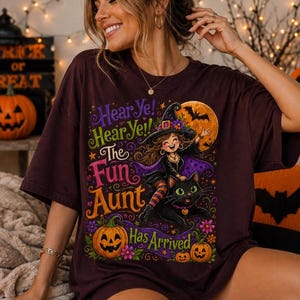 K&ouml;nnte beinhalten: Ein dunkelbraunes T-Shirt mit einer Grafik einer l&auml;chelnden Hexe, die auf einer schwarzen Katze reitet. Der Text lautet Hear Ye! Hear Ye! The Fun Aunt Has Arrived. Das Design zeigt einen gro&szlig;en orangefarbenen Mond, eine Fledermaus, zwei K&uuml;rbisse und Blumen.