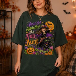 K&ouml;nnte beinhalten: Ein dunkelgr&uuml;nes T-Shirt mit einer bunten Grafik einer Cartoon-Hexe, die auf einer schwarzen Katze reitet. Der Text lautet "Hear Ye! Hear Ye! The Fun Aunt Has Arrived" in Lila, Gr&uuml;n und Orange. Das Design enth&auml;lt einen gelben Mond und orange K&uuml;rbisse.