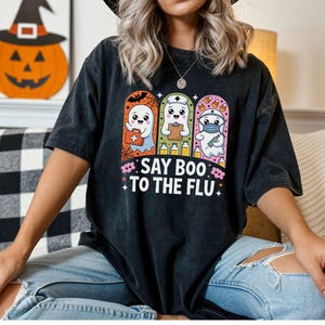 May include: Black t-shirt featuring a Halloween-themed graphic with three cartoon ghosts dressed as a doctor, nurse, and first responder. The text "SAY BOO TO THE FLU" is below the ghosts. A pumpkin with a witch hat is in the background.
