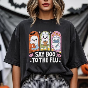 May include: Black t-shirt with a Halloween-themed graphic. The design features three cartoon ghosts dressed as a doctor, nurse, and medic, with the text "SAY BOO TO THE FLU". The ghosts are set against a backdrop of bats and pumpkins.