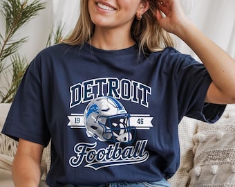 Op de afbeelding: Marineblauw T-shirt met de woorden "DETROIT Football" in witte letters en blokletters. Een zilveren en blauwe voetbalhelm is gecentreerd onder het woord "DETROIT". Het shirt heeft ook de nummers "19" en "46".