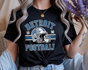 Op de afbeelding: Zwart T-shirt met een Detroit Lions football ontwerp. Het ontwerp bevat het woord "DETROIT" in blauw, de jaartallen "19" en "30", een footballhelm en het woord "FOOTBALL".