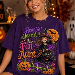 K&ouml;nnte beinhalten: Ein violettes T-Shirt mit einer bunten Grafik einer Hexe, die auf einer schwarzen Katze reitet. Der Text lautet: Hear Ye! Hear Ye! The Fun Aunt Has Arrived. Das Design zeigt einen gro&szlig;en orangefarbenen Mond, Flederm&auml;use und K&uuml;rbisse.
