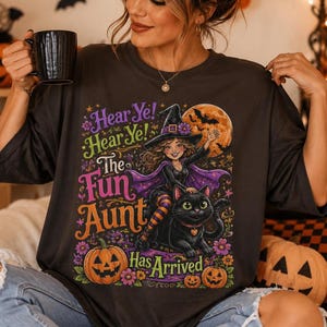 K&ouml;nnte beinhalten: Ein dunkelgraues T-Shirt mit einer bunten Grafik einer Hexe, die auf einer schwarzen Katze reitet. Der Text lautet: Hear Ye! Hear Ye! The Fun Aunt Has Arrived. Das Design zeigt einen gro&szlig;en orangefarbenen Mond, Flederm&auml;use, Blumen und zwei K&uuml;rbisse.