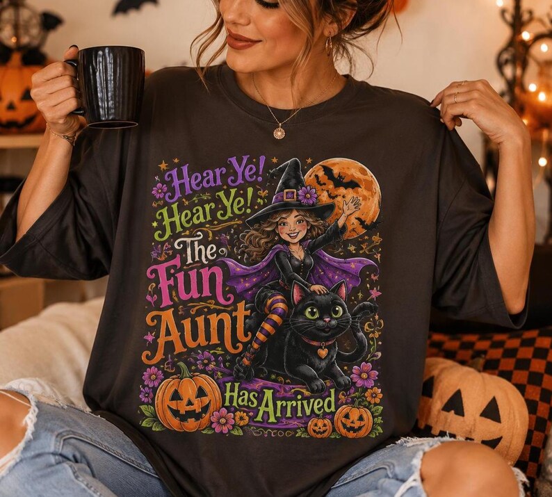 K&ouml;nnte beinhalten: Ein dunkelgraues T-Shirt mit einer bunten Grafik einer Hexe, die auf einer schwarzen Katze reitet. Der Text lautet: Hear Ye! Hear Ye! The Fun Aunt Has Arrived. Das Design zeigt einen gro&szlig;en orangefarbenen Mond, Flederm&auml;use, Blumen und zwei K&uuml;rbisse.