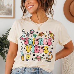 Pode incluir: Uma t-shirt de cor creme com o texto "SCIENCE VIBES" numa fonte colorida de estilo retro. O design inclui várias ilustrações de temática científica, como um foguetão, planetas, um microscópio, uma lâmpada, um tubo de ensaio e um béquer.
