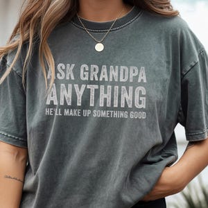 May include: Dark gray t-shirt with the text "ASK GRANDPA ANYTHING, HE'LL MAKE UP SOMETHING GOOD" in white, distressed lettering. The shirt has a vintage, washed look and a relaxed fit, suitable for casual wear.