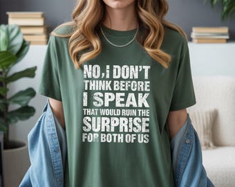 May include: A dark green t-shirt with the text "NO, I DON'T THINK BEFORE I SPEAK THAT WOULD RUIN THE SURPRISE FOR BOTH OF US" in white block letters. The shirt is worn with a denim jacket.