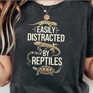 May include: Charcoal grey t-shirt with the slogan "EASILY DISTRACTED BY REPTILES" in white. The design includes illustrations of a lizard, a snake, and a turtle in brown and green tones.