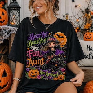 K&ouml;nnte beinhalten: Ein schwarzes T-Shirt mit einer bunten Grafik einer Hexe, die auf einer schwarzen Katze reitet, mit dem Text Hear Ye! Hear Ye! The Fun Aunt Has Arrived. Das Design enth&auml;lt einen gro&szlig;en orangefarbenen Mond, lila und orange Blumen und einen K&uuml;rbis.