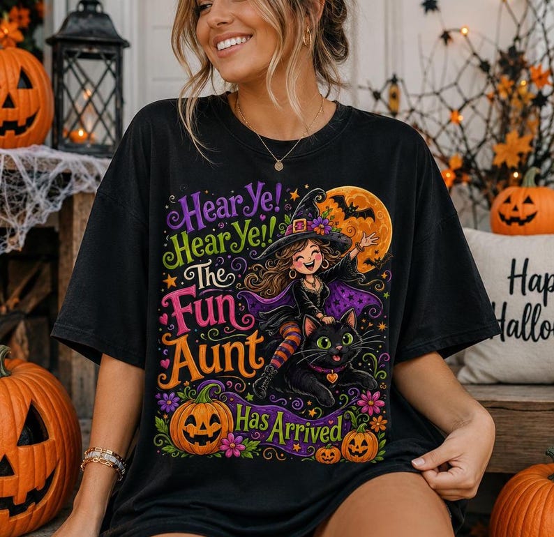 K&ouml;nnte beinhalten: Ein schwarzes T-Shirt mit einer bunten Grafik einer Hexe, die auf einer schwarzen Katze reitet, mit dem Text Hear Ye! Hear Ye! The Fun Aunt Has Arrived. Das Design enth&auml;lt einen gro&szlig;en orangefarbenen Mond, lila und orange Blumen und einen K&uuml;rbis.