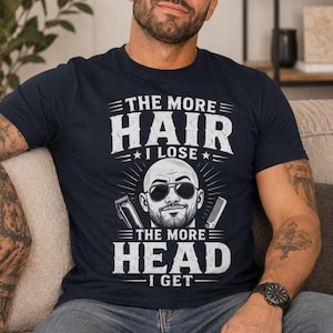 May include: A navy blue short-sleeve t-shirt featuring a white graphic of a bald person wearing sunglasses, flanked by a hair clipper and a comb. The text reads: THE MORE HAIR I LOSE THE MORE HEAD I GET, arranged in a bold, distressed font style.