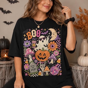 May include: A black t-shirt featuring a colorful graphic print with the text BOO and STAY SPOOKY. The design includes a ghost wearing heart-shaped glasses, a jack-o'-lantern, mushrooms, a crescent moon, a disco ball, and various floral elements.