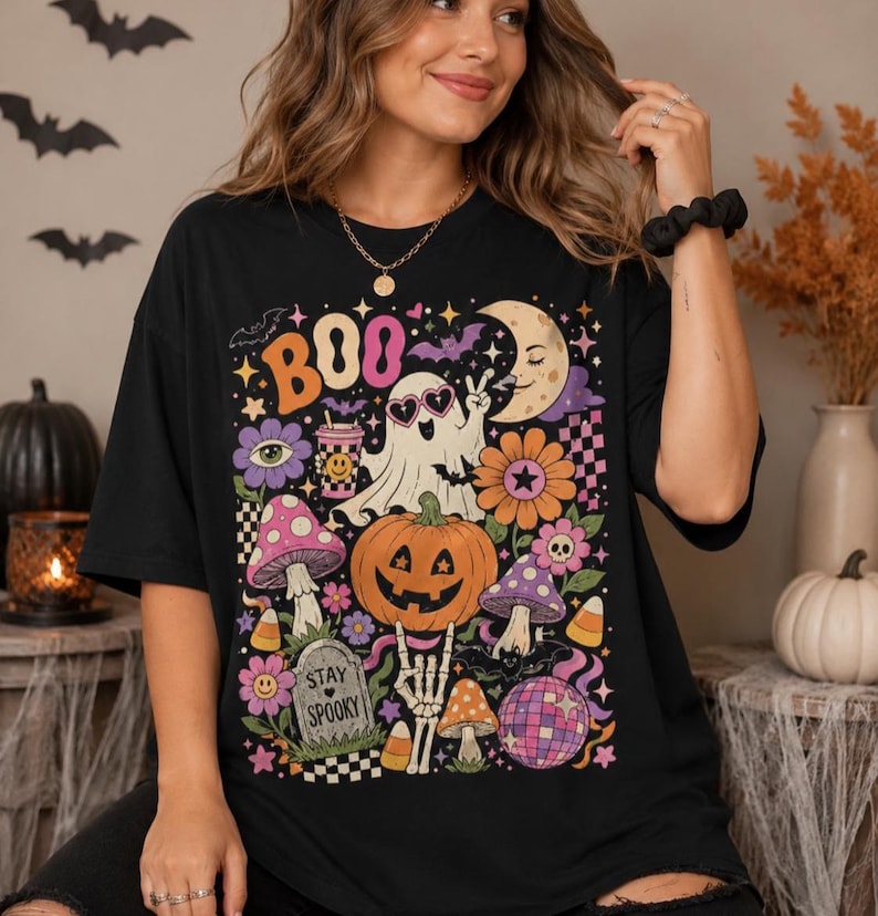 May include: A black t-shirt featuring a colorful graphic print with the text BOO and STAY SPOOKY. The design includes a ghost wearing heart-shaped glasses, a jack-o'-lantern, mushrooms, a crescent moon, a disco ball, and various floral elements.