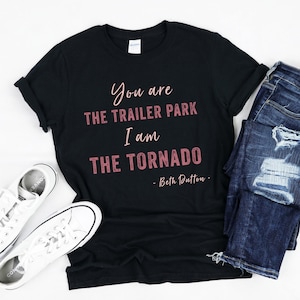 Könnte beinhalten: Schwarzes T-Shirt mit dem Zitat "You are the Trailer Park, I am the Tornado - Beth Dutton" in rosa Schrift. Das Shirt wird mit weißen Turnschuhen und einer blauen Jeans kombiniert.