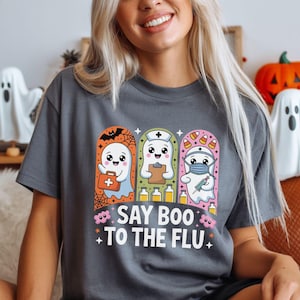 May include: Gray t-shirt with a Halloween-themed graphic. The graphic features three cartoon ghosts dressed as a doctor, nurse, and medic, with the text "Say Boo to the Flu". Decorative ghost figurines and a pumpkin are in the background.