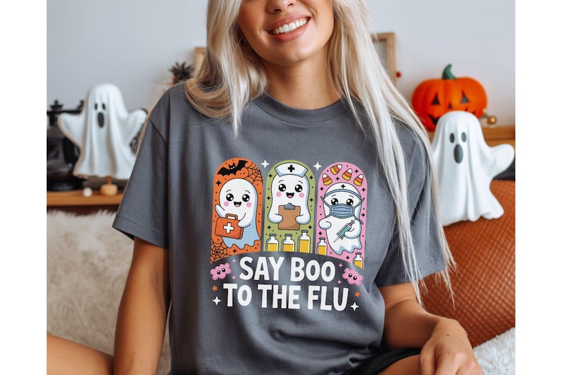 May include: Gray t-shirt with a Halloween-themed graphic. The graphic features three cartoon ghosts dressed as a doctor, nurse, and medic, with the text "Say Boo to the Flu". Decorative ghost figurines and a pumpkin are in the background.