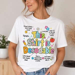 Op de afbeelding: Een wit t-shirt met korte mouwen en een kleurrijke afbeelding met de tekst This Shirt is Decodable. Het ontwerp bevat educatieve iconen zoals een potlood, appel en alfabetblokken, omringd door taalkundige aantekeningen over lettergrepen.