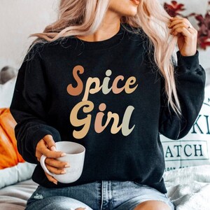 Peut inclure: Sweat-shirt noir avec l'inscription "Spice Girl" dans une police rétro. Le texte est dans des tons de marron, pêche et vert. La personne sur l'image tient une tasse blanche. Le sweat-shirt est de style décontracté.