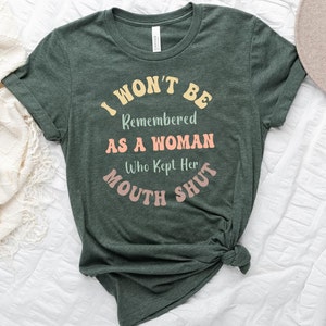 Puede incluir: Una camiseta verde oscuro con el texto "I won't be remembered as a woman who kept her mouth shut" en una fuente retro. El texto está en amarillo, rosa y naranja.