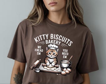 May include: Brown t-shirt with a graphic of a cat wearing a chef's hat, kneading dough. The text reads "Kitty Biscuits Bakery" with the phrases "We Knead Em" and "You Need Em". A rolling pin, bowl, and biscuits are also shown.