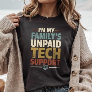 Może przedstawiać: Czarny t-shirt z napisem "I'M MY FAMILY'S UNPAID TECH SUPPORT" w stylu retro. Tekst jest w kolorze turkusowym, żółtym i czerwonym. Mała grafika komputera znajduje się na dole. Osoba nosi beżowy kardigan.