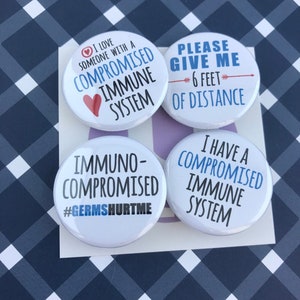 May include: Four white round buttons with text. One says "I LOVE SOMEONE WITH A COMPROMISED IMMUNE SYSTEM" with a heart. Another says "PLEASE GIVE ME 6 FEET OF DISTANCE". The third says "IMMUNO-COMPROMISED #GERMSHURTME". The last says "I HAVE A COMPROMISED IMMUNE SYSTEM".