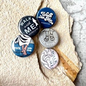 Könnte beinhalten: Eine Sammlung von fünf runden Buttons mit dem Satz "Walk with Me" und zugehörigen Bildern. Die Buttons zeigen verschiedene Designs, darunter Silhouetten, Sandalen und stilisierte Wellen, mit dem Text "Moses 6:34". Die Buttons sind in Blau-, Schwarz-, Weiß- und Rosatönen gehalten.