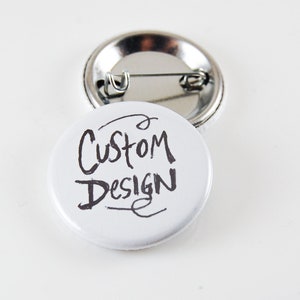 May include: Two white round buttons with a pin back. One button has the words "Custom Design" in black handwritten style. The other button is visible in the background.