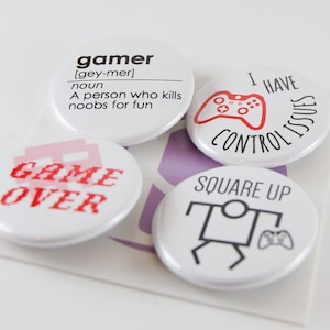 May include: Four white round buttons with gaming-related text and graphics. One button defines "gamer", another says "Game Over" in red pixelated font, a third says "I have control issues", and the last says "Square Up".
