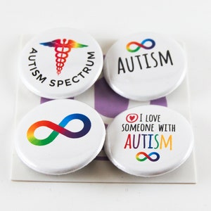 May include: Four white round buttons with autism-related designs. One button has a rainbow caduceus and the words "AUTISM SPECTRUM". Another has a rainbow infinity symbol and the word "AUTISM". The third has a rainbow infinity symbol. The fourth says "I LOVE SOMEONE WITH AUTISM".