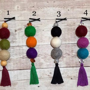 May include: Four colorful felt ball garlands with wooden beads and tassels. Each garland has a different color scheme, including red, green, orange, purple, black, white, gray, teal, and pink.