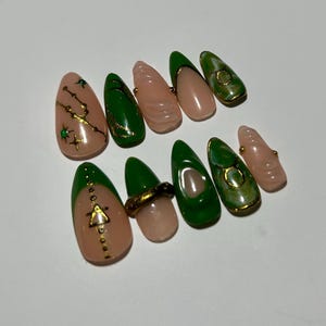 May include: A set of ten artificial nails in various designs. Some nails are a light pink with gold accents, while others are a vibrant green with gold trim. The nails feature star and geometric patterns.