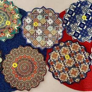 May include: Seven colorful round coasters with intricate mandala and geometric patterns. The coasters are made of a hard material and have a slightly raised edge. They are arranged in a circle on a red and blue mat.