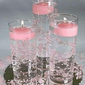 Può includere: Tre vasi cilindrici in vetro trasparente con candele galleggianti rosa e acqua limpida con piccole perle bianche. I vasi sono su una superficie nera con perle bianche sparse intorno.