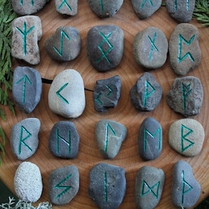 Puede incluir: Conjunto de 24 piedras grises con runas verdes pintadas en ellas. Las piedras están dispuestas en círculo sobre una superficie de madera.