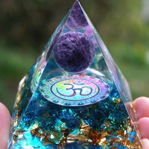 Puede incluir: Una pirámide de cristal transparente con una esfera de amatista púrpura en su interior. La pirámide está llena de cristales azules y verdes y escamas doradas. El símbolo "Om" está impreso en la base de la pirámide.